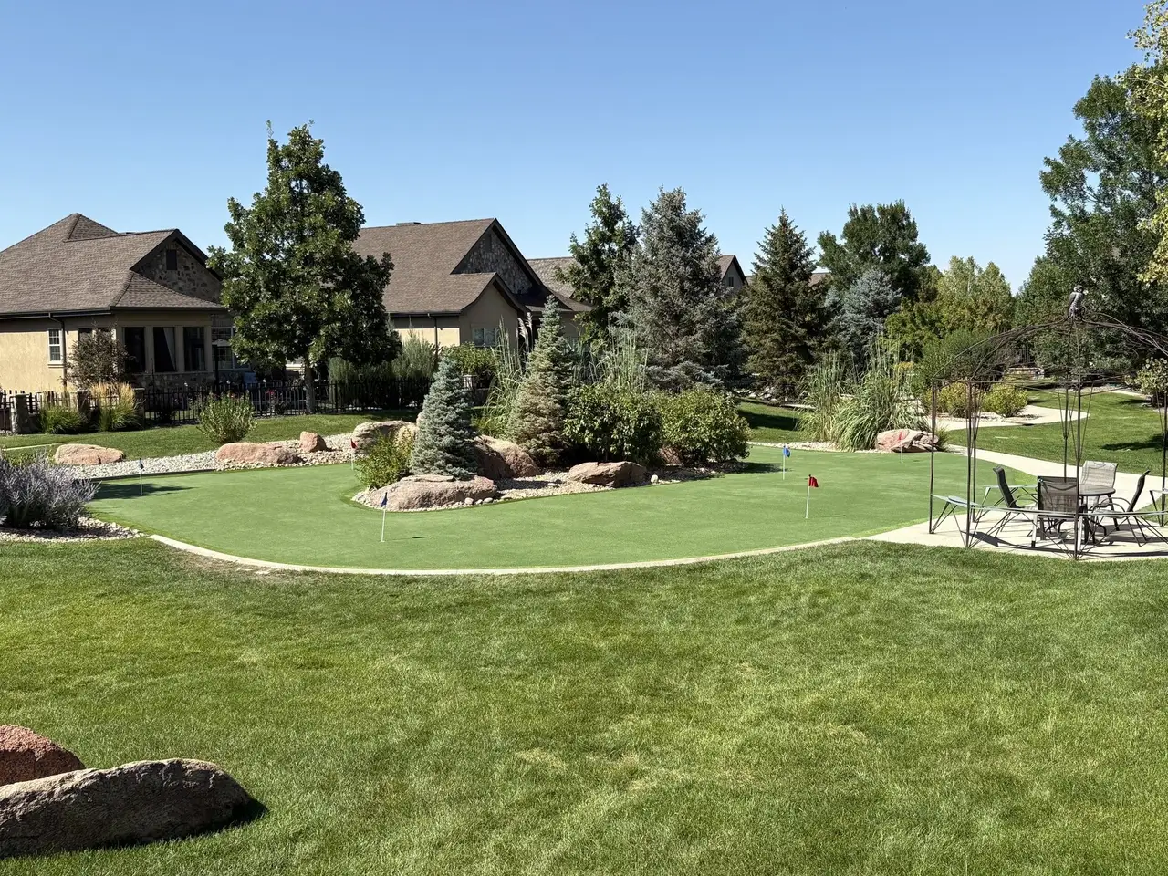 A backyard putting green ringed with boulders and planting beds, flag in the cup, house behind