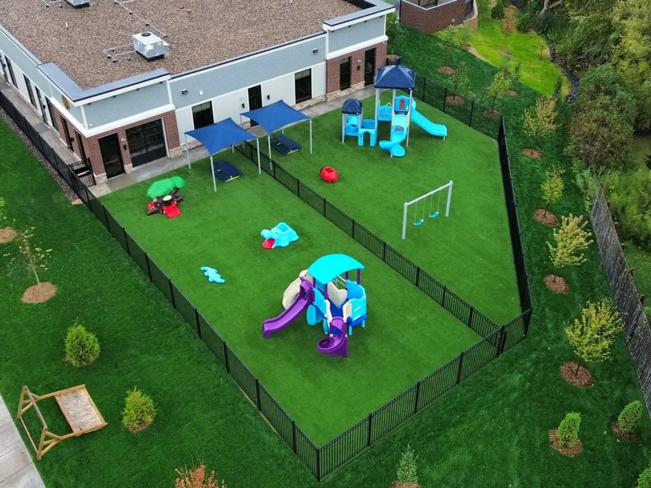 An aerial view of a fenced play yard at a child care building, turf under the play structures, swings and shade sails