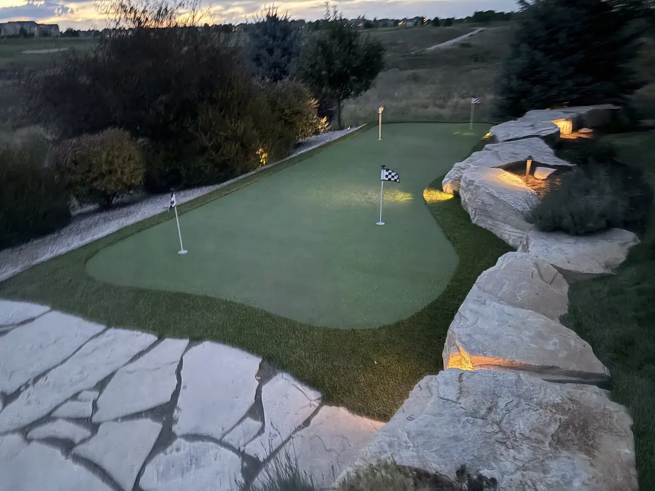 A putting green at dusk with landscape lighting on the flagstone and two flags in