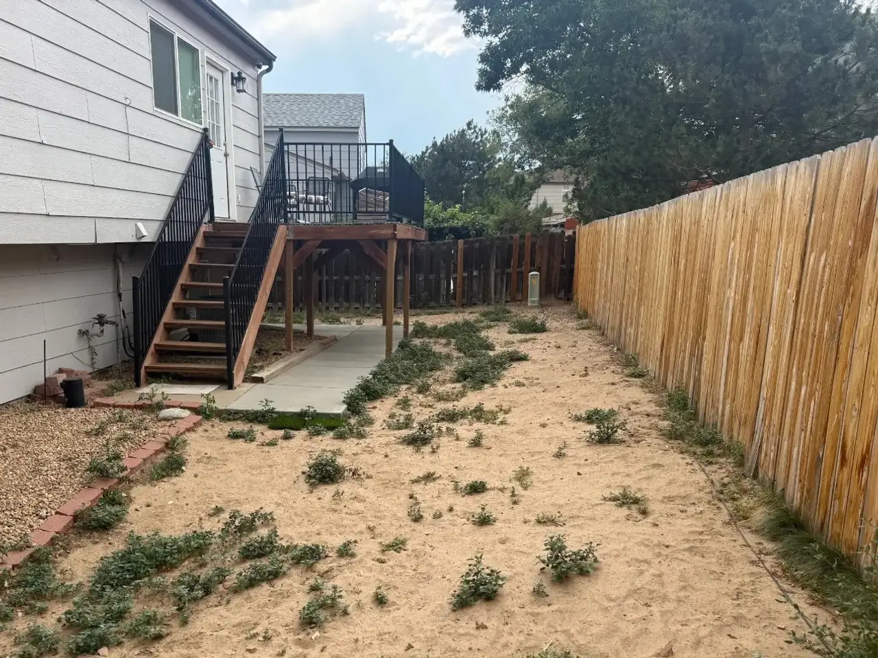 A fenced side yard of bare sand and weeds, with a gravel bed and brick edging along the house and deck stairs behind